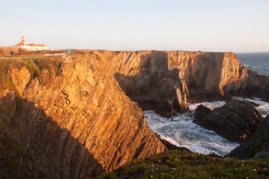 Kayalıklarla kaplı engebeli kıyı manzarası, sörf dalgaları, denizdeki kayalar, yumuşak güneş ışığıyla aydınlatılmış.