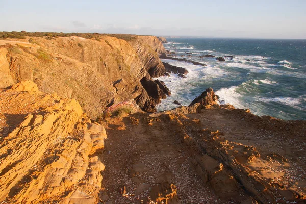 Kayalıklarla kaplı engebeli kıyı manzarası, sörf dalgaları, denizdeki kayalar, yumuşak güneş ışığıyla aydınlatılmış.