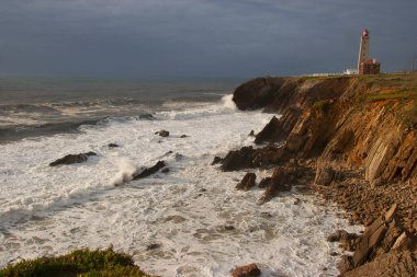 Kayalıklarla kaplı engebeli kıyı manzarası, sörf dalgaları, denizdeki kayalar, yumuşak güneş ışığıyla aydınlatılmış.