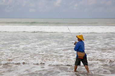 Sahilde şapkalı bir adam balık tutuyor.