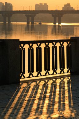 Dnipro Nehri yakınlarındaki Kyiv 'de sonbahar gündoğumu manzarası. Klasik metal çitler kaldırım taşlarının üzerine gölge düşürür. Arka planda Dinyeper Nehri üzerindeki ünlü Paton Köprüsü 'nün bulanık silueti.