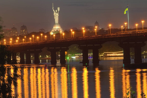Autumn night cityscape with Paton bridge over Dnieper river. Motherland monument at the background. City lights reflected in the water. Kyiv, Ukraine.