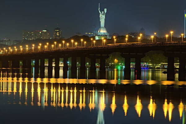 Autumn night cityscape with Paton bridge over Dnieper river. Motherland monument at the background. City lights reflected in the water. Kyiv, Ukraine.
