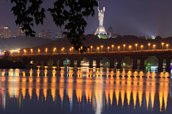 Motherland Monument at the top of the hill in tree leaves border. Natural frame. Autumn night panorama of Dnipro River, skyscrapers in the background.  City lights reflected in the water.Kyiv,Ukraine.