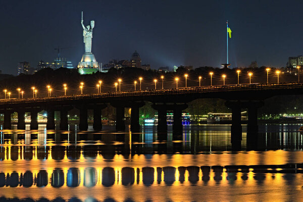 Night cityscape panorama of illuminated Paton bridge over Dnieper river. Famous Motherland monument at the background. City lights reflected in the water. Kyiv, Ukraine.