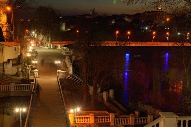 Kamianets-Podilskyi şehir parkının gece manzarası. Yürüyen insanların bulanık siluetleri. Ünlü turistik mekan ve romantik seyahat merkezi. Arka planda Smotrych Nehri üzerindeki köprü..