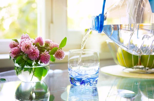 Blue glass beaker, water is poured from a large plastic bottle. In the kitchen near the window  on the table glass of water and a vase of flowers.
