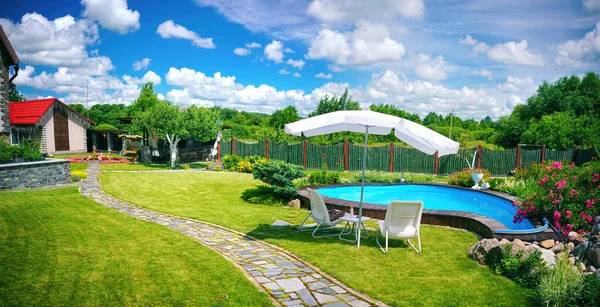 Small pool and garden furniture in backyard with beautiful manicured lawn and flower bed on warm sunny summer day against backdrop of blue sky with clouds.