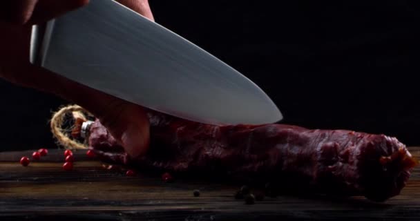 Mens hand with a knife cuts in half a loaf of sausage lying on a wooden board — Stock Video