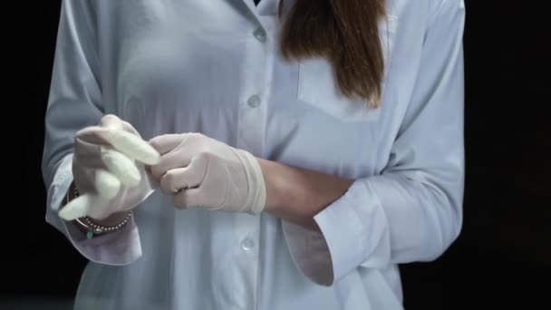 Junge Ärztin in weißer Robe auf schwarzem Hintergrund legt Latex-Handschuhe an ihre Hände — Stockvideo