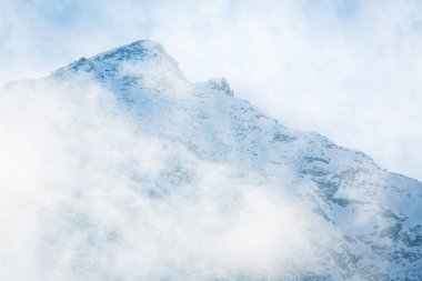 Kış karıyla kaplı görkemli bir dağ zirvesi kısmen sürüklenen sis ve alçak bulutlarla gizlenmiş. Huzurlu yüksek irtifa manzarası doğa, macera, kış yolculuğu ve dağ keşifleri için mükemmeldir..
