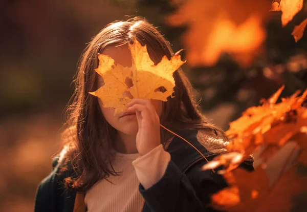 A 10-year-old girl walks in the park in a green coat in autumn.Yellow leaves, a beautiful girl on a walk. Long hair, a smile. Warm orange background.