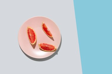 Üç parça taze greyfurt dilimi pastel pembe tabakta gri, mavi arka planda. Belgelerle birlikte en az yaz plajı konsepti. Kopyalama alanı olan düz.
