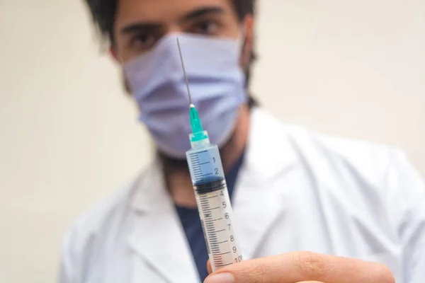 İçinde mavi sıvı aşı bulunan plastik şırınga parmaklarıyla odaklanmış, beyaz önlüklü ve maskeli bir doktor aşıya hazır, arka planda çözülmüş. COVID-19 Coronavirüs salgını