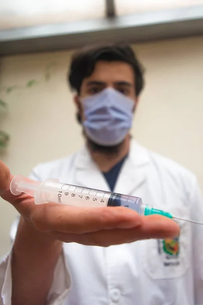 İçinde mavi sıvı aşı bulunan plastik şırınga parmaklarıyla odaklanmış, beyaz önlüklü ve maskeli bir doktor aşıya hazır, arka planda çözülmüş. COVID-19 Coronavirüs salgını