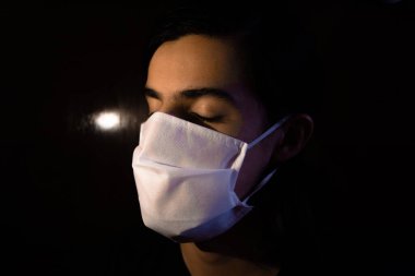 Tek kullanımlık beyaz maskeli bir adamın portresi. Gözleri kapalı ve ciddi bir ifadeyle. Siyah arkaplanlı gösterişsiz ışık fotoğrafçılığı. COVID Coronavirus salgını sahnesi