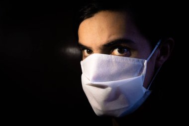 Tek kullanımlık beyaz maskeli bir adamın portresi kameraya ciddi bir ifadeyle bakıyor. Siyah arkaplanlı gösterişsiz ışık fotoğrafçılığı. COVID Coronavirus salgını sahnesi