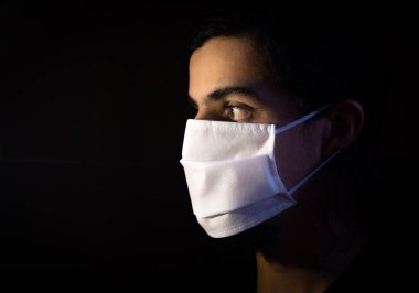 Tek kullanımlık beyaz maskeli bir adamın portresi kameraya ciddi bir ifadeyle bakıyor. Siyah arkaplanlı gösterişsiz ışık fotoğrafçılığı. COVID Coronavirus salgını sahnesi