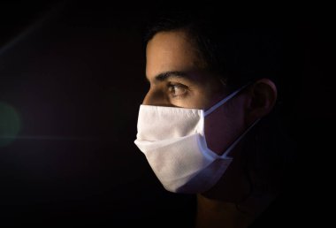 Tek kullanımlık beyaz maskeli bir adamın portresi kameraya ciddi bir ifadeyle bakıyor. Siyah arkaplanlı gösterişsiz ışık fotoğrafçılığı. COVID Coronavirus salgını sahnesi