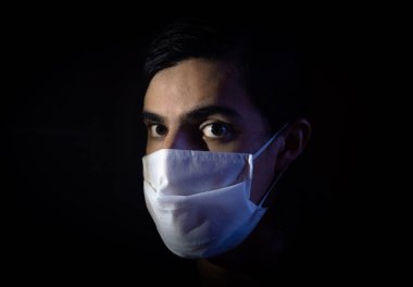 Tek kullanımlık beyaz maskeli bir adamın portresi. Sürpriz bir ifadeyle kameraya bakıyor. Siyah arkaplanlı gösterişsiz ışık fotoğrafçılığı. COVID-19 veya koronavirüs salgını sahnesi