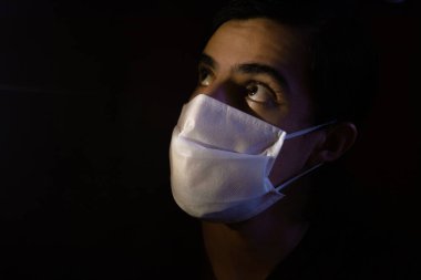 Tek kullanımlık beyaz maskeli bir adamın portresi. Sürpriz bir ifadeyle kameraya bakıyor. Siyah arkaplanlı gösterişsiz ışık fotoğrafçılığı. COVID-19 veya koronavirüs salgını sahnesi