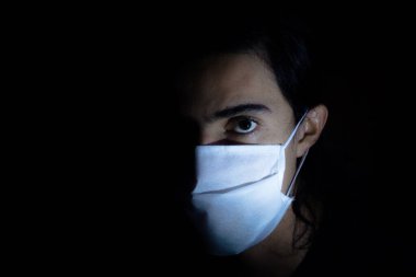 Tek kullanımlık beyaz maskeli bir adamın portresi kameraya ciddi bir ifadeyle bakıyor. Siyah arkaplanlı gösterişsiz ışık fotoğrafçılığı. COVID Coronavirus salgını sahnesi