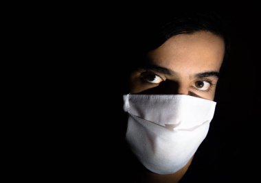 Tek kullanımlık beyaz maskeli bir adamın portresi kameraya ciddi bir ifadeyle bakıyor. Siyah arkaplanlı gösterişsiz ışık fotoğrafçılığı. COVID Coronavirus salgını sahnesi