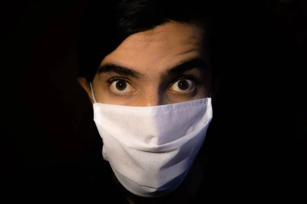 Tek kullanımlık beyaz maskeli bir adamın portresi. Sürpriz bir ifadeyle kameraya bakıyor. Siyah arkaplanlı gösterişsiz ışık fotoğrafçılığı. COVID-19 veya koronavirüs salgını sahnesi
