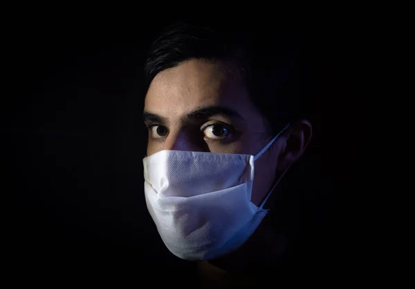 Portrait of a man with a disposable white face mask looking at the camera with a surprise expression face. Low key light photography with black background. COVID-19 or coronavirus pandemic scene