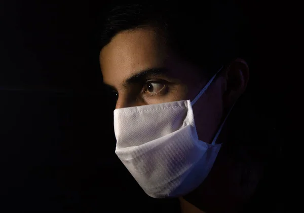 Tek kullanımlık beyaz maskeli bir adamın portresi kameraya ciddi bir ifadeyle bakıyor. Siyah arkaplanlı gösterişsiz ışık fotoğrafçılığı. COVID Coronavirus salgını sahnesi