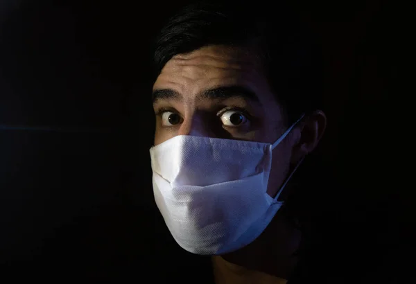 Tek kullanımlık beyaz maskeli bir adamın portresi. Sürpriz bir ifadeyle kameraya bakıyor. Siyah arkaplanlı gösterişsiz ışık fotoğrafçılığı. COVID-19 veya koronavirüs salgını sahnesi