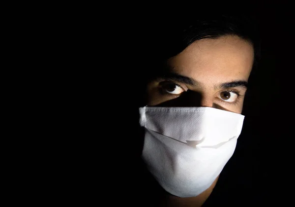 Tek kullanımlık beyaz maskeli bir adamın portresi kameraya ciddi bir ifadeyle bakıyor. Siyah arkaplanlı gösterişsiz ışık fotoğrafçılığı. COVID Coronavirus salgını sahnesi