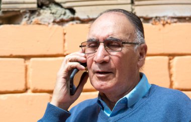 Gözlüklü yaşlı bir adamın portresi arka planda turuncu bir duvarla dışarıda telefonla konuşuyor. COVID veya Coronavirus salgını sahnesi.