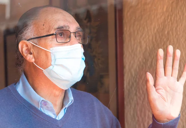 Gözlüklü ve tıbbi maskeli yaşlı bir adamın portresi, camların arkasında, gökyüzünün ve binaların yansıması olan bir camın arkasına bakıyordu. COVID veya Coronavirus salgını sahnesi.