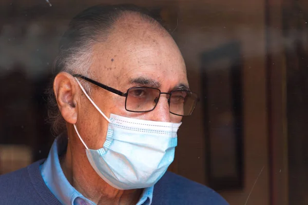 Portrait od an old senior man with glasses and medical face mask looking outside behind a glass with reflection of buildings and sky, hand on the glass. Scene of COVID or Coronavirus pandemic.