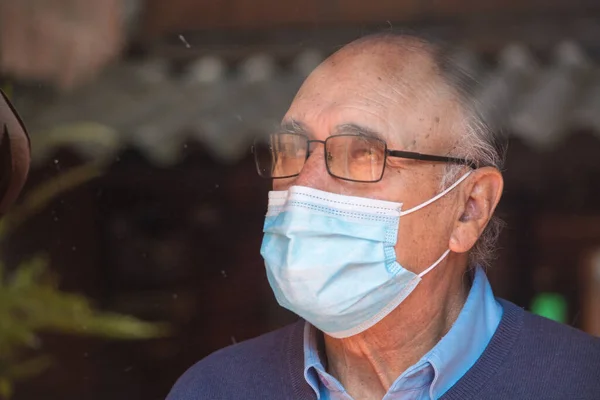 Gözlüklü ve tıbbi maskeli yaşlı bir adamın portresi, camların arkasında, gökyüzünün ve binaların yansıması olan bir camın arkasına bakıyordu. COVID veya Coronavirus salgını sahnesi.
