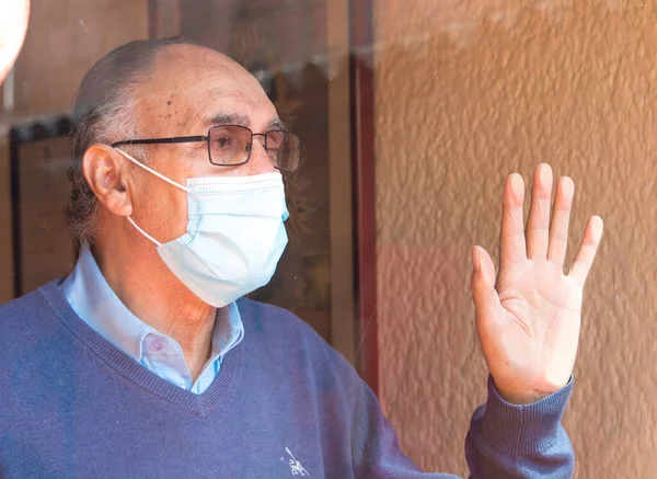 Gözlüklü ve tıbbi maskeli yaşlı bir adamın portresi, camların arkasında, gökyüzünün ve binaların yansıması olan bir camın arkasına bakıyordu. COVID veya Coronavirus salgını sahnesi.