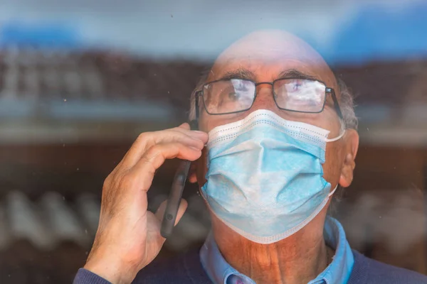 Gözlüklü ve tıbbi maskeli yaşlı bir adamın portresi, camların ve gökyüzünün yansıması olan bir camın arkasında telefonla konuşuyor. COVID veya Coronavirus salgını sahnesi.