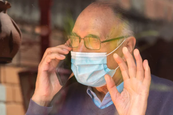 Portrait od an old senior man with glasses and medical face mask talking on the phone behind a glass with reflection of buildings and sky, hand on the glass. Scene of COVID or Coronavirus pandemic.