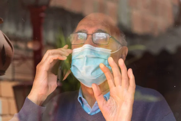 Portrait od an old senior man with glasses and medical face mask talking on the phone behind a glass with reflection of buildings and sky, hand on the glass. Scene of COVID or Coronavirus pandemic.