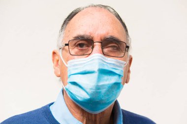 Gözlüklü yaşlı bir doktorun portresi ve mavi bir tıbbi yüz maskesi ve kameraya bakan mavi bir süveter. Beyaz arka plan. Covid veya koronavirüs salgını sahnesi.