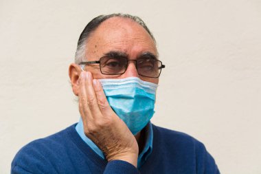 Gözlük takan yaşlı bir doktorun portresi ve tıbbi yüz maskesi, endişeli bir şekilde kameraya bakıyor. COVID 19 ya da Coronavirus salgınının sahnesi.