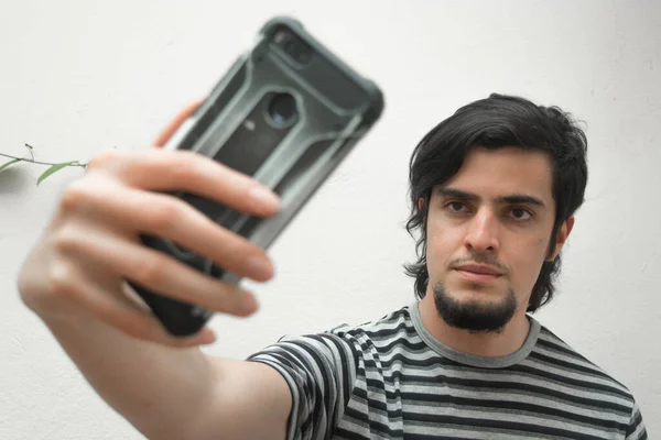 Elinde telefon tutan sakallı bir gencin portresi ciddi ve yakışıklı bir ifadeyle selfie çekiyor. Siyah beyaz tişört giyen bir manken.. 