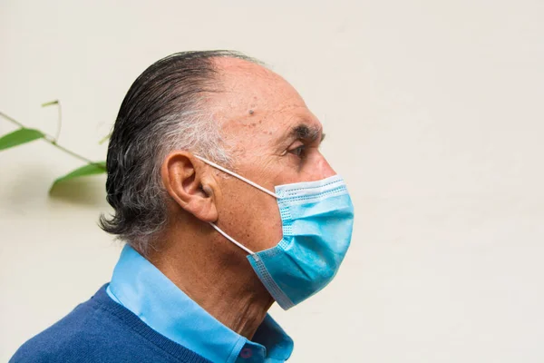 Profile portrait of a doctor senior old man wearing glasses and a blue medical face mask, and a blue sweater, man looking at camera. White background. Scene of covid or coronavirus pandemic.