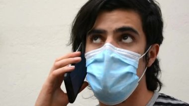 Yüz maskesi ve çizgili gömlekli beyaz bir adam akıllı bir telefonla konuşuyor ya da telefonla konuşuyor. Siyah saç modeli, beyaz arkaplan