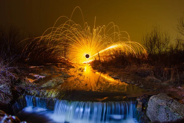 Искры из горящей стальной шерсти, отраженные в ручье возле водопада — стоковое фото