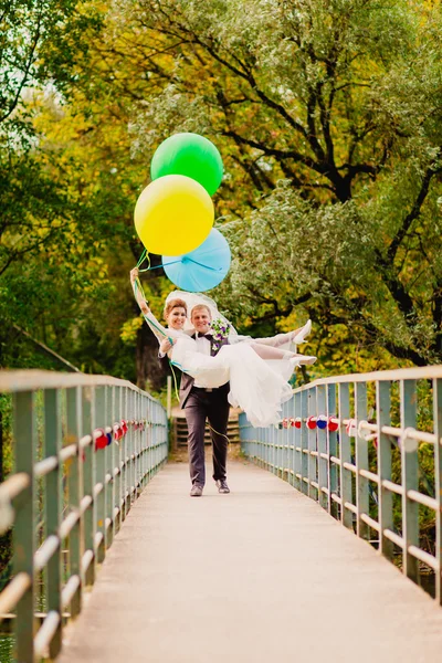 Der Bräutigam trägt seine Braut mit Luftballons über die Brücke — Stockfoto