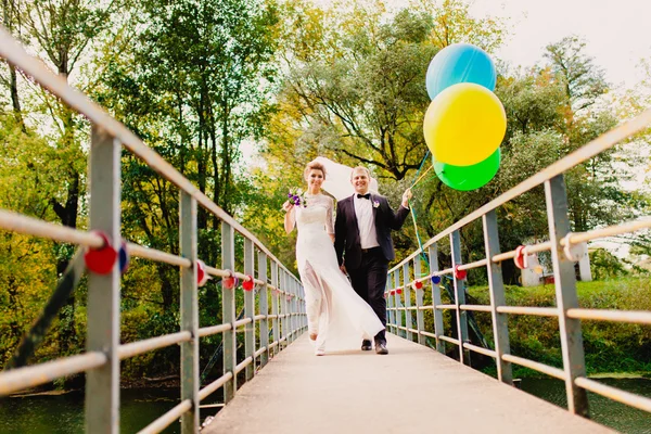 Braut und Bräutigam gehen auf der Brücke mit Luftballons vor dem Hintergrund der herbstlichen Landschaft — Stockfoto