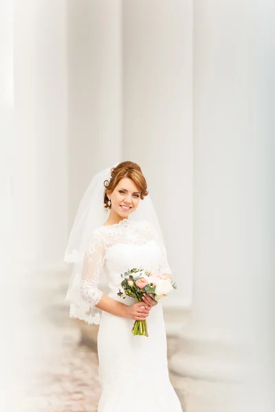 Ritratto di una bella giovane sposa che si erge tra le colonne e regge un bouquet da sposa — Foto Stock