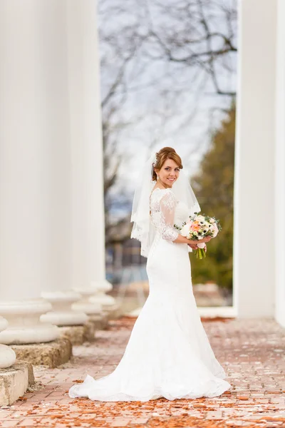 Giovane bella sposa in piedi tra i pilastri e in possesso di un bouquet da sposa — Foto Stock